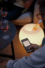 Ein beleuchteter, kugelförmiger Tisch mit einer Flasche Wein, umgeben von Personen, die ein Smartphone benutzen. Perspektive von oben.