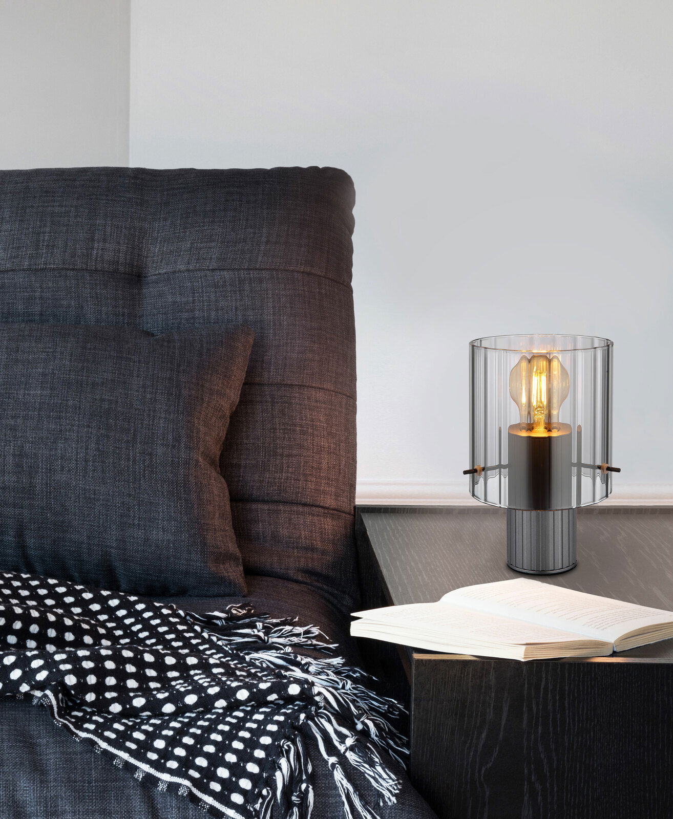 Moderne Tischleuchte TULIVAR mit transparentem Glasschirm auf einem Nachttisch neben einem gemütlichen Sessel, seitliche Perspektive.
