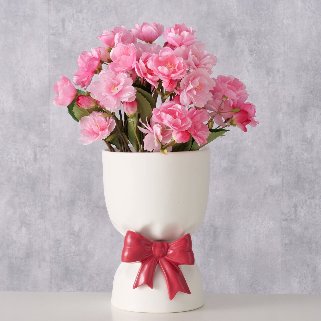 Weiße Porzellanvase Bowy, 22 cm hoch, mit roter Schleife und rosa Blumen, frontal fotografiert.