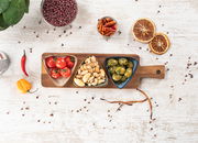 Vogelperspektive auf ein Akazienbrett mit drei dreieckigen Schalen, gefüllt mit Tomaten, Nüssen und Oliven, umgeben von Gewürzen und getrockneten Orangenscheiben auf einem hellen Holztisch.