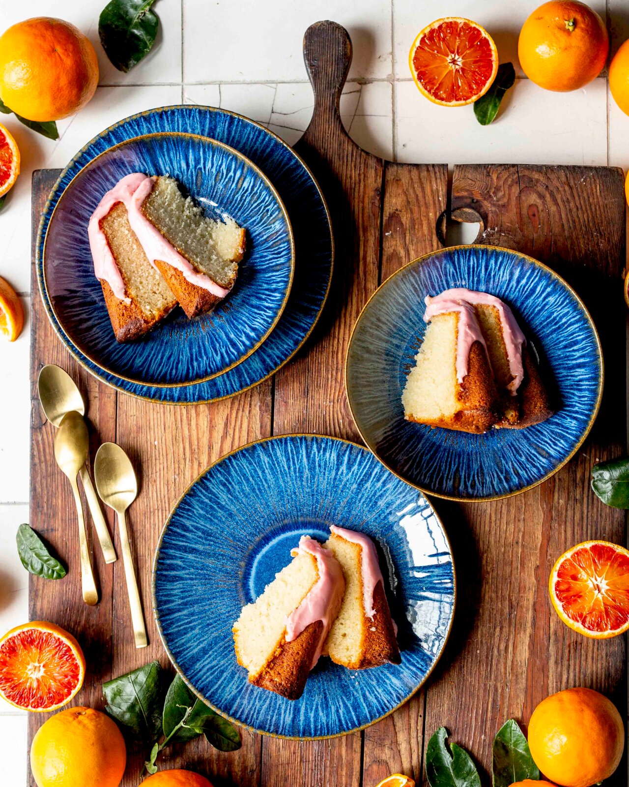 Drei blaue Keramikteller mit Stücken von Rührkuchen mit rosa Glasur auf rustikalem Holzbrett, umgeben von Orangen und grünen Blättern, Draufsicht (Flatlay)