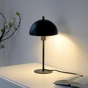 Schwarze, minimalistische Tischlampe mit halbkugelförmigem Schirm auf weißem Tisch, eingeschaltet mit warmem Lichtkegel an der Wand, Frontansicht in leichter Seitenperspektive.