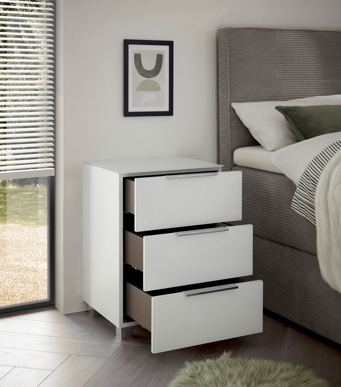 Moderne weiße Nachtkommode mit drei offenen Schubladen, seitliche Perspektive neben einem Bett.