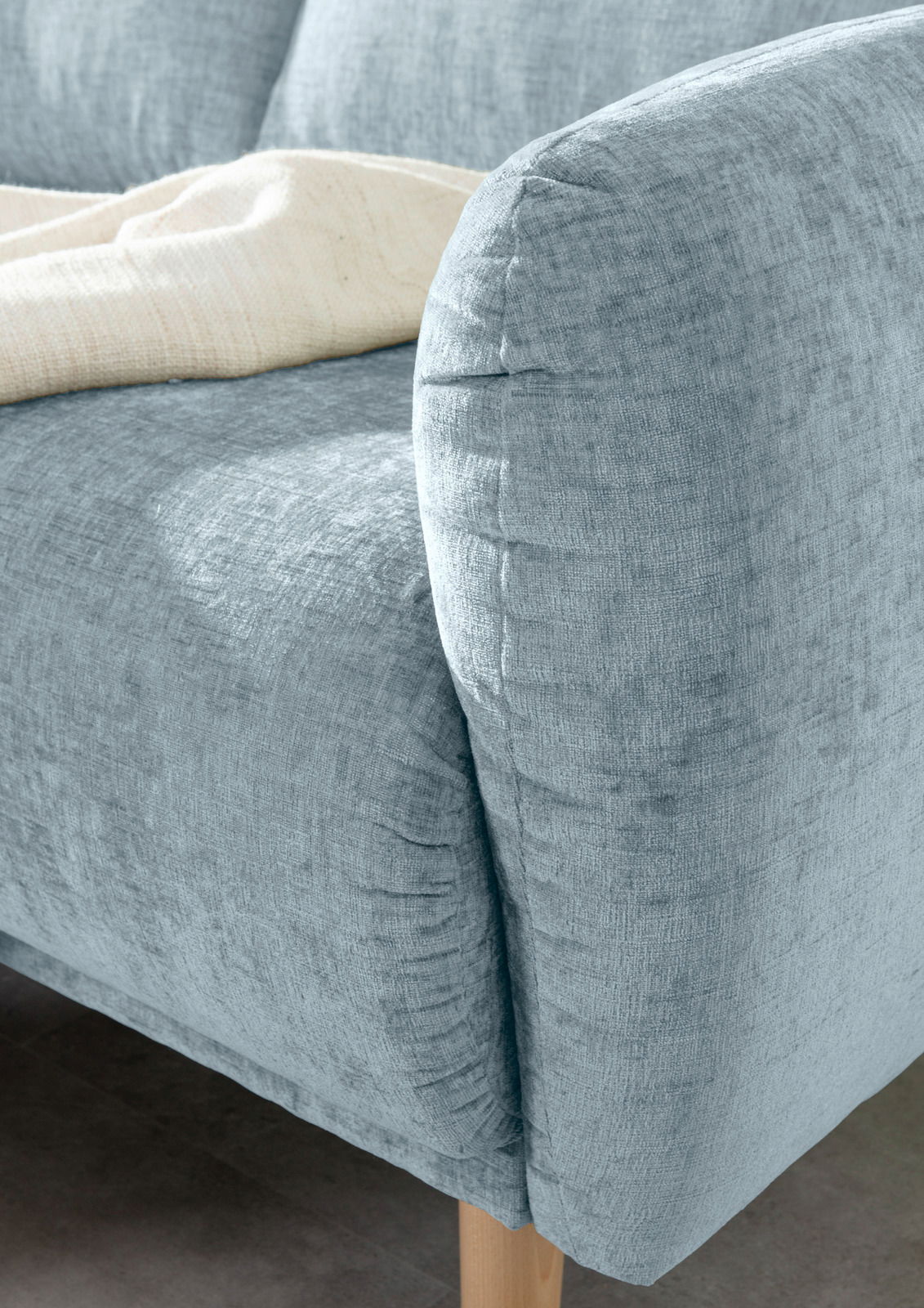Nahaufnahme einer hellblauen Sofaecke mit abgerundeter Armlehne und Holzfuß, von der Seite betrachtet.