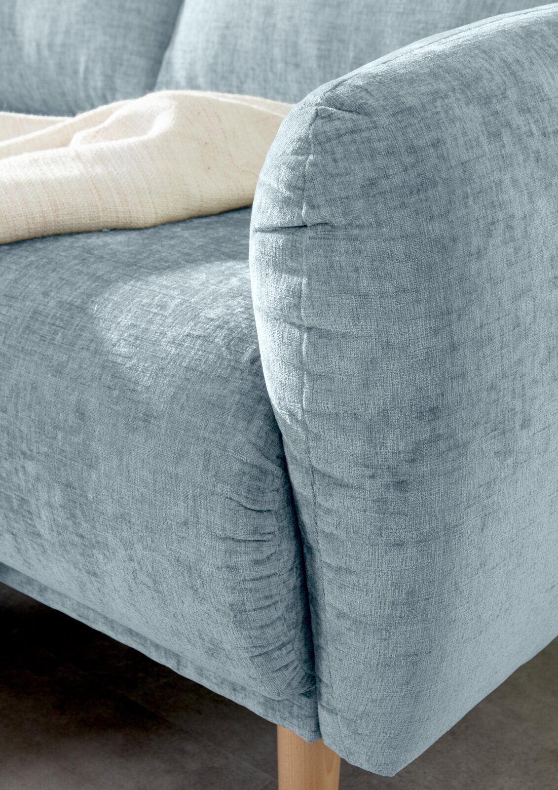 Nahaufnahme einer hellblauen Sofaecke mit abgerundeter Armlehne und Holzfuß, von der Seite betrachtet.