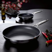 Zwei schwarze Pfannen mit Edelstahlgriffen auf dunklem Tisch, vorn eine große Bratpfanne und hinten eine kleinere Pfanne, seitliche Perspektive mit leicht erhöhter Ansicht