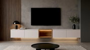 Frontale Ansicht eines modernen, schwebenden TV-Lowboards in Weiß mit mittigem Holzfach und indirekter LED-Beleuchtung unter einem wandmontierten Flachbildfernseher; links Tischlampe und Bücher, rechts Vase mit Zweigen.