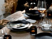 Elegant gedeckter Esstisch mit schwarzem Geschirr, goldenem Besteck, geriffelten Wein- und Wassergläsern sowie stimmungsvollen Kerzen, aus leicht erhöhter, schräger Vorderansicht fotografiert.