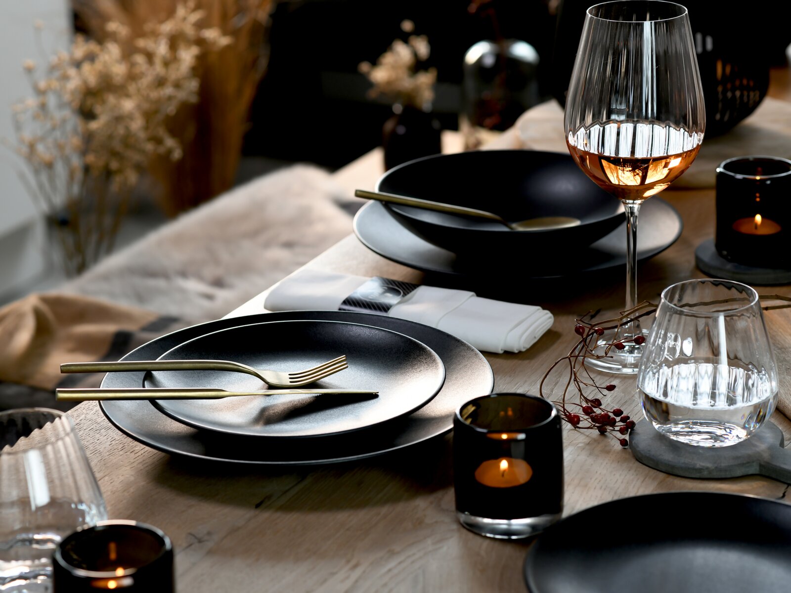 Elegant gedeckter Esstisch mit schwarzem Geschirr, goldenem Besteck, geriffelten Wein- und Wassergläsern sowie stimmungsvollen Kerzen, aus leicht erhöhter, schräger Vorderansicht fotografiert.