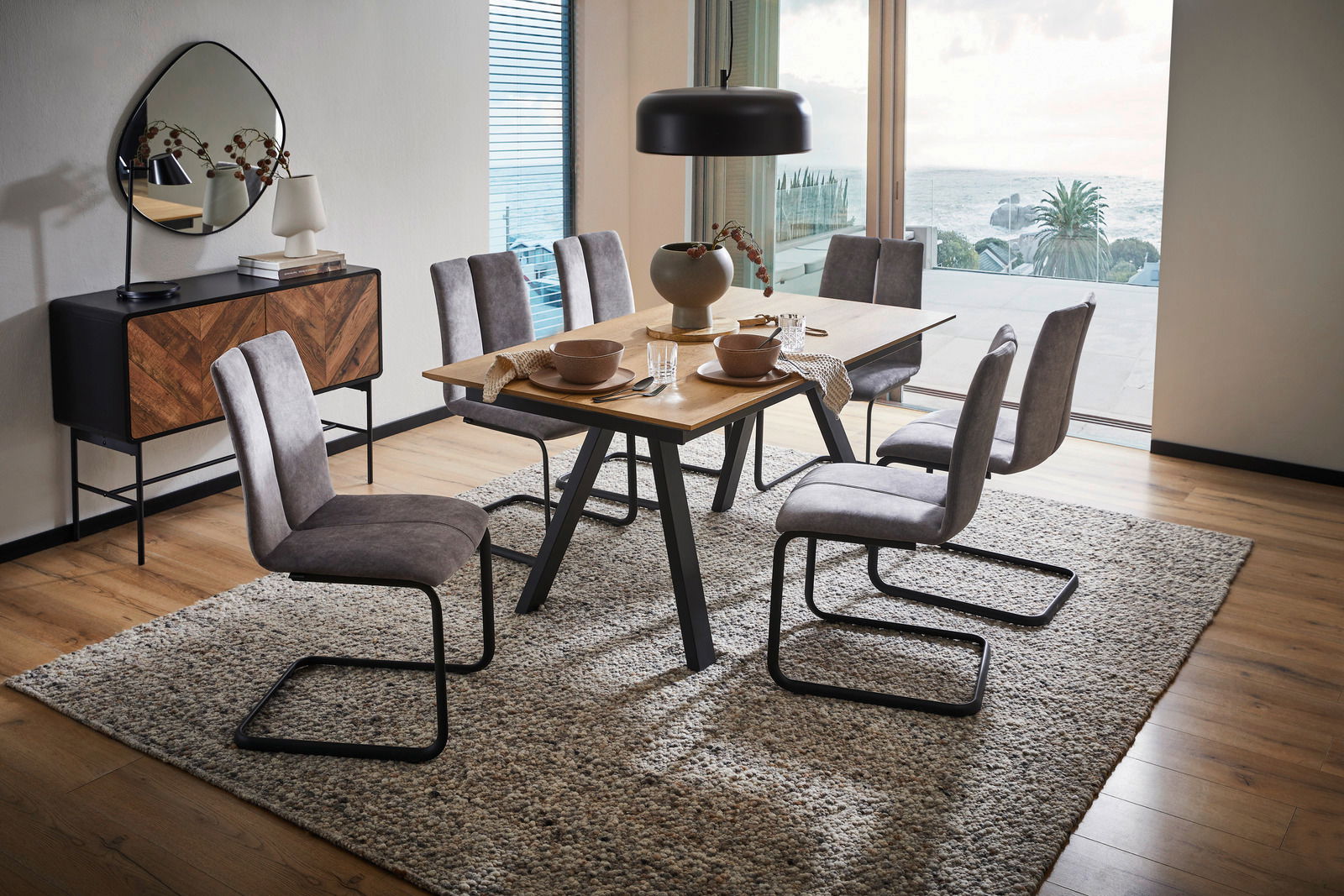 Moderner Esstisch mit Holzplatte und schwarzen Metallbeinen, umgeben von sechs grauen Stühlen auf einem Teppich, mit Blick auf eine große Fensterfront mit Meerblick.