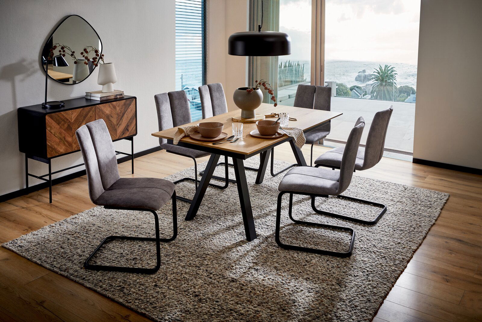 Moderner Esstisch mit Holzplatte und schwarzen Metallbeinen, umgeben von sechs grauen Stühlen auf einem Teppich, mit Blick auf eine große Fensterfront mit Meerblick.