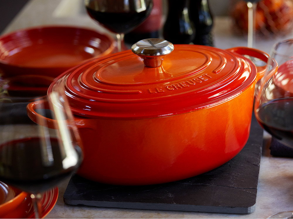 Roter Le Creuset Bräter auf einem gedeckten Tisch, hinter gefüllten Rotwein Gläsern.