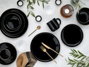 Draufsicht auf ein elegant gedecktes Tischarrangement mit glänzend schwarzem Geschirr mit feinem Goldrand, schwarzer Tasse, goldfarbenem Besteck, Kerzen, Wassergläsern und grünen Zweigen auf hellem Hintergrund.