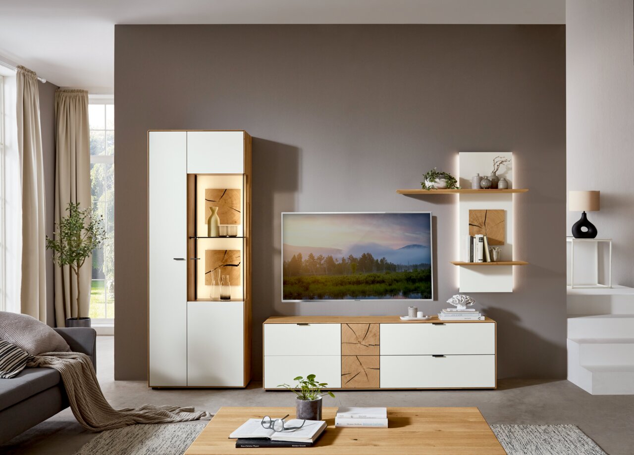 Moderne Wandkombination von Wöstmann Markenmöbel, bestehend aus einem hohen Schrank mit Glaselementen, einem TV-Board und zwei Wandregalen, in einer stilvollen Wohnzimmerumgebung. Frontalansicht.