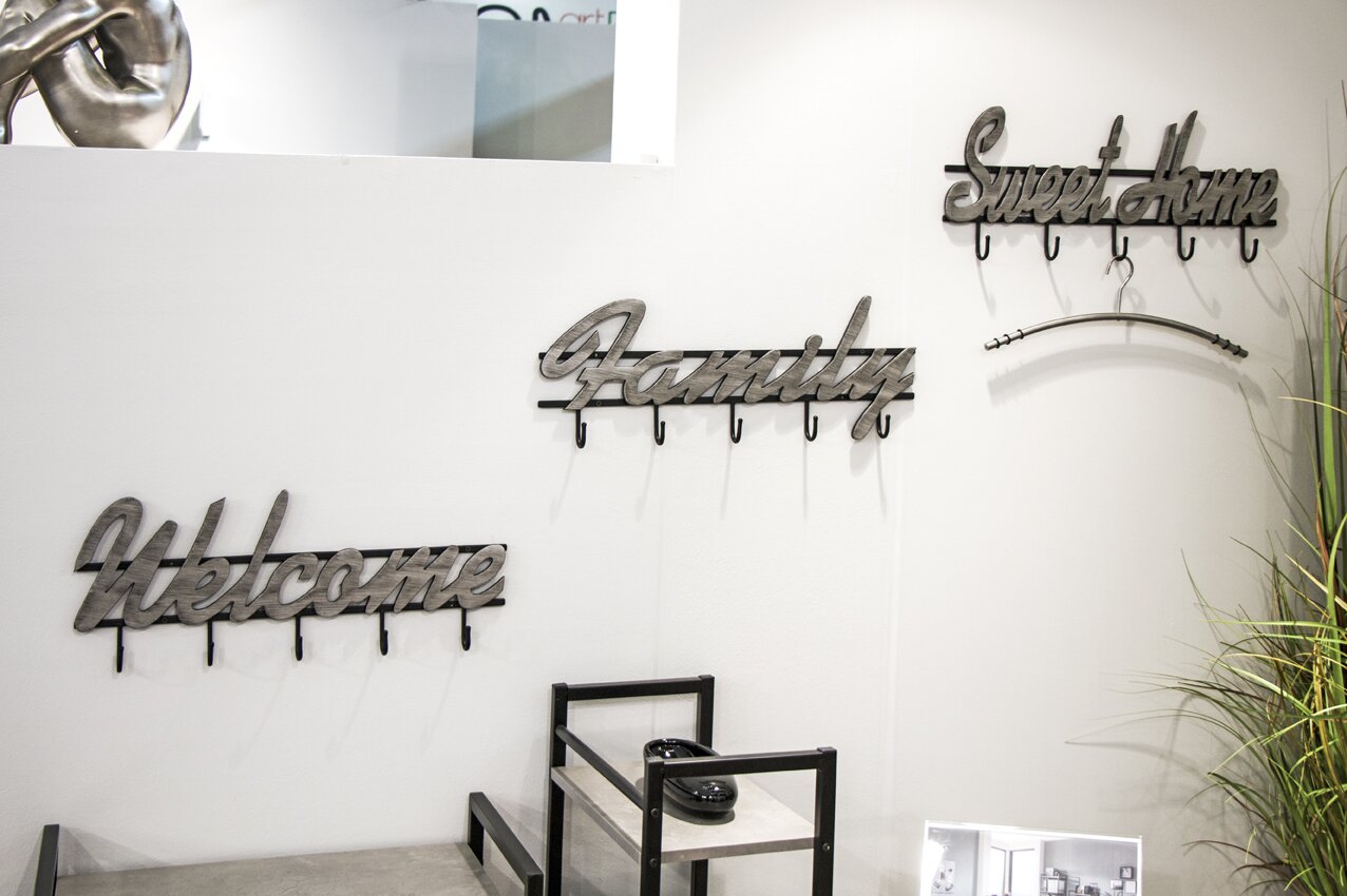 Frontalansicht einer weißen Wand mit dekorativen Wandgarderoben in Schriftzug-Optik („Welcome“, „Family“, „Sweet home“) aus Metall/Holzoptik mit mehreren Haken, darüber ein Regal und rechts eine Pflanze.