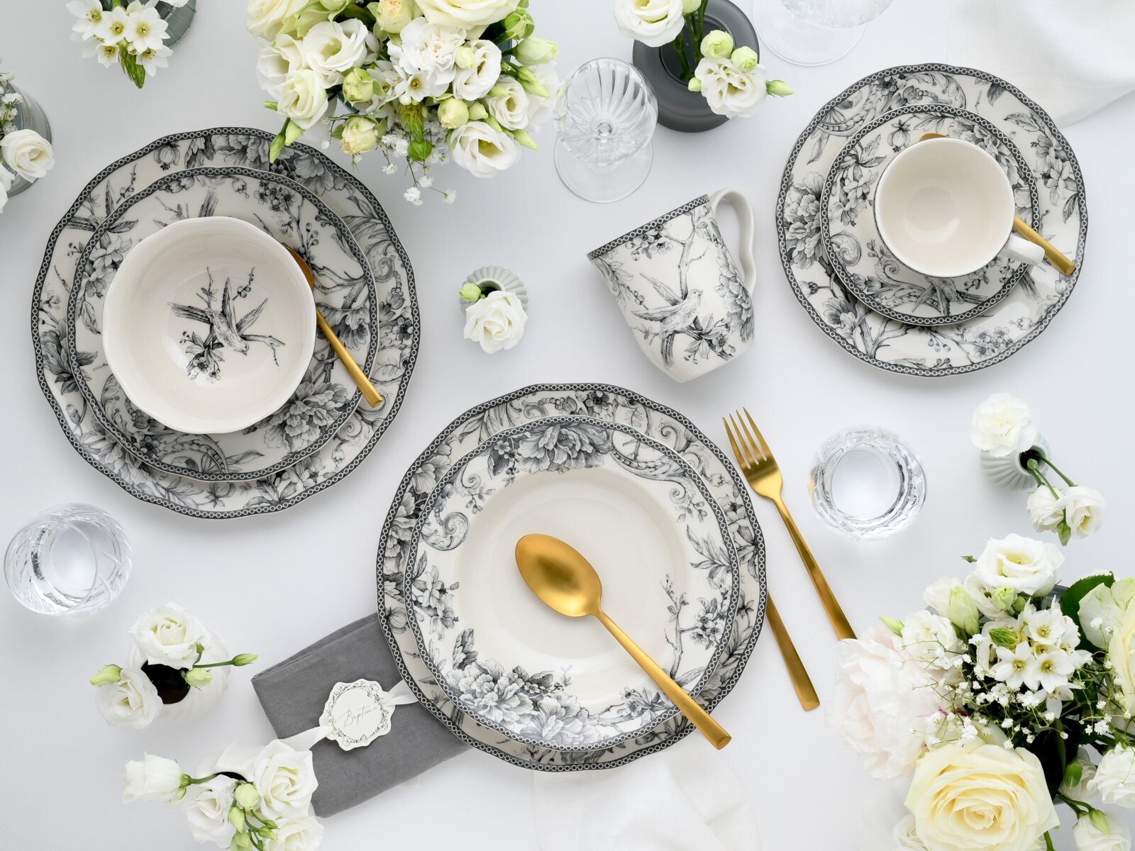 Elegantes schwarz-weißes Porzellan-Geschirrset mit floralen Motiven (Teller, Schalen und Tasse) mit goldfarbenem Besteck und weißen Blumen auf weißem Tisch, Draufsicht von oben