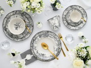 Elegantes schwarz-weißes Porzellan-Geschirrset mit floralen Motiven (Teller, Schalen und Tasse) mit goldfarbenem Besteck und weißen Blumen auf weißem Tisch, Draufsicht von oben