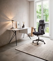 Moderner Schreibtisch mit schlanken Metallbeinen und Schubladen, seitliche Perspektive, neben einem ergonomischen Bürostuhl in einem hellen Raum mit großem Fenster.