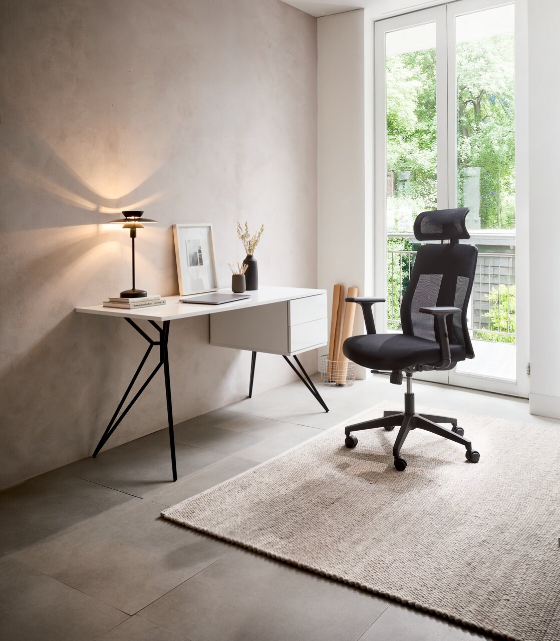 Moderner Schreibtisch mit schlanken Metallbeinen und Schubladen, seitliche Perspektive, neben einem ergonomischen Bürostuhl in einem hellen Raum mit großem Fenster.