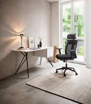 Moderner Schreibtisch mit schlanken Metallbeinen und Schubladen, seitliche Perspektive, neben einem ergonomischen Bürostuhl in einem hellen Raum mit großem Fenster.