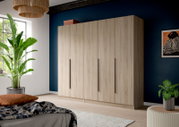 Großer Kleiderschrank in heller Holzoptik mit vier Türen und langen schwarzen Griffen, in einem Schlafzimmer vor dunkelblauer Wand, perspektivisch in leichter Schrägansicht von vorn rechts.