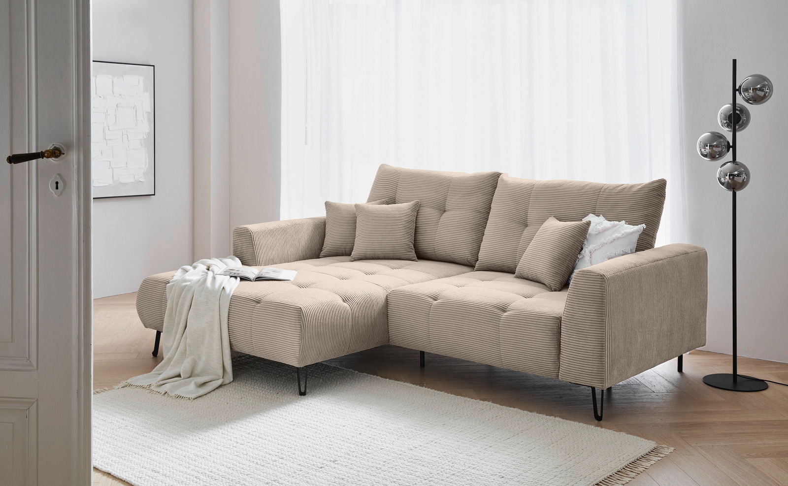 Beige Ecksofa mit Ottomane links, dekoriert mit Kissen und Decke, in einem modernen Wohnzimmer mit hellem Holzboden und Stehlampe. Perspektive von der Seite.