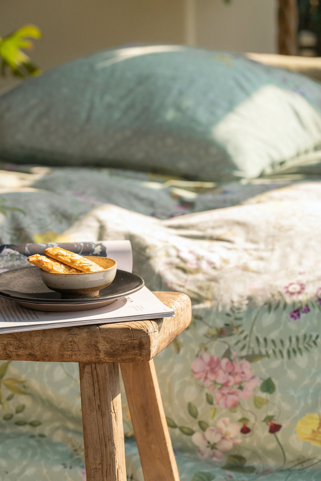 Nahaufnahme einer Bettwäsche mit floralem Muster in sanften Farben, im Vordergrund ein rustikaler Holztisch mit einer Schale und Gebäck, seitliche Perspektive.