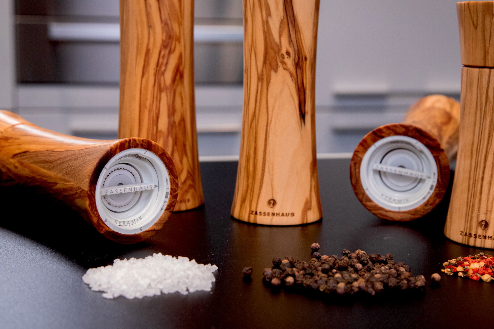 Salzmühle aus Olivenholz in Nahaufnahme, liegend und stehend, mit Salz und Pfefferkörnern im Vordergrund.
