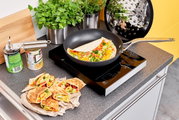 Een koekenpan van Raise met een diameter van 20 cm op een inductiekookplaat, gevuld met een kleurrijke omelet, omringd door verse kruiden en ingrediënten, gefotografeerd van bovenaf.