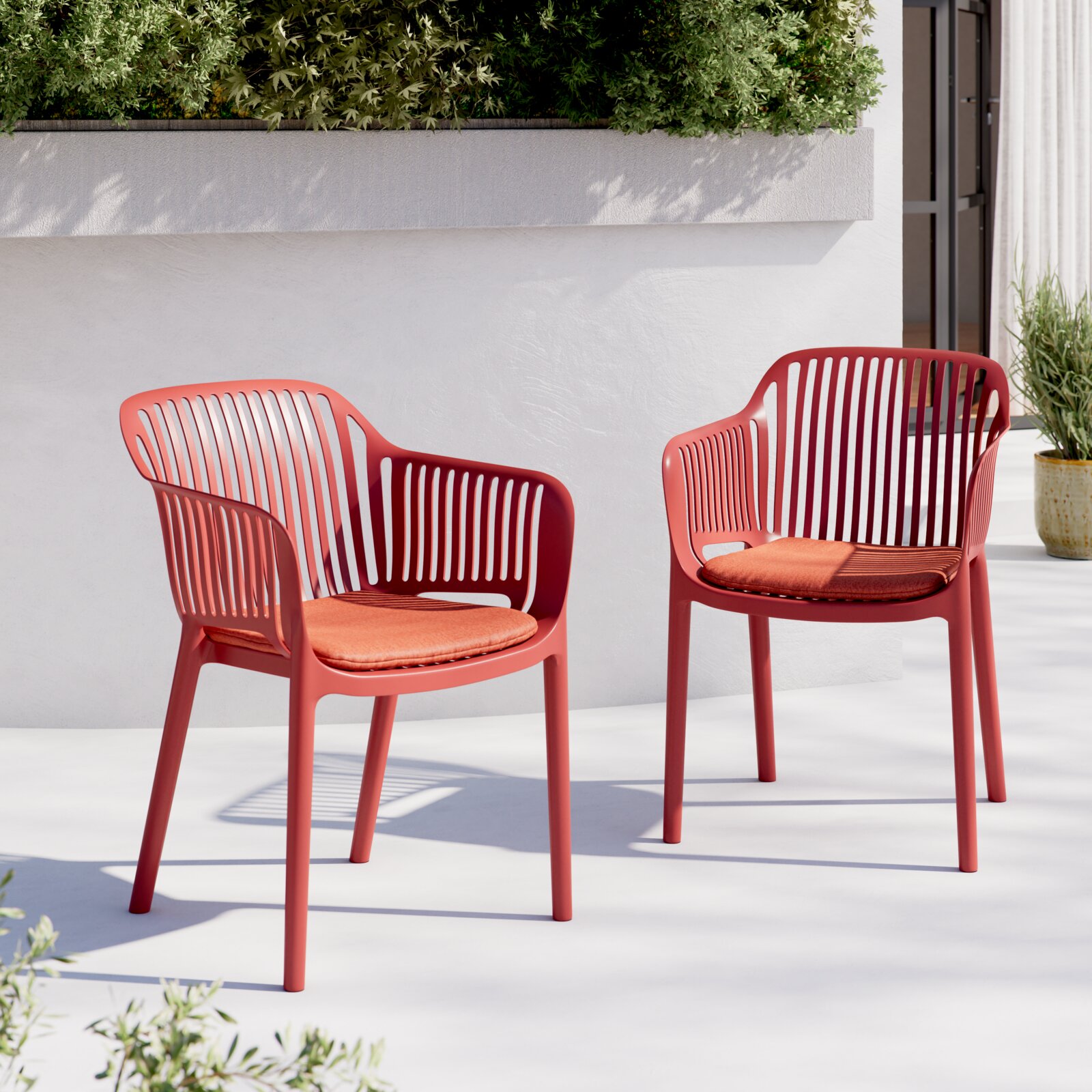 Twee moderne rode kunststof tuinstoelen met oranje zitkussens op een lichtgekleurd terras voor een witte muur en een plantenbak, perspectief: licht schuin van voren op ooghoogte