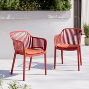 Twee moderne rode kunststof tuinstoelen met oranje zitkussens op een lichtgekleurd terras voor een witte muur en een plantenbak, perspectief: licht schuin van voren op ooghoogte