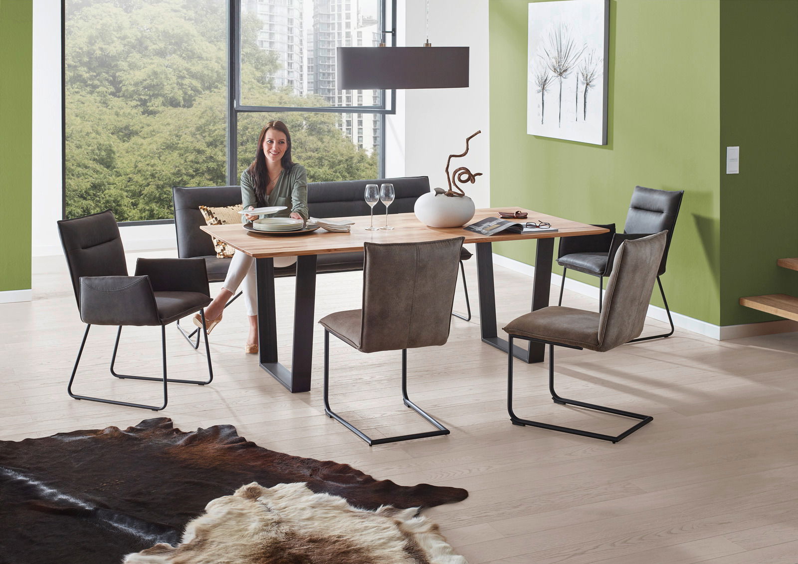 Valnatura Sessel SCALA Moderner Essbereich mit Esstisch aus Holz und Metallgestell, umgeben von Freischwinger-Stühlen und einer Bank, in einem hellen Raum mit großen Fenstern und grüner Wand. Perspektive: seitlich.