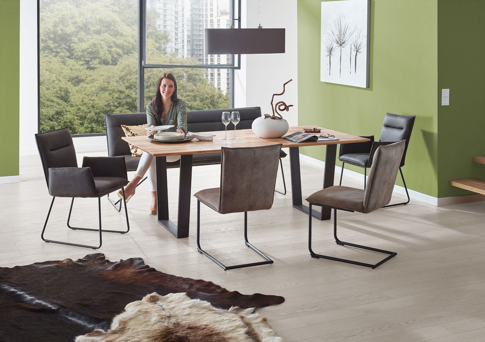 Valnatura Sessel SCALA Moderner Essbereich mit Esstisch aus Holz und Metallgestell, umgeben von Freischwinger-Stühlen und einer Bank, in einem hellen Raum mit großen Fenstern und grüner Wand. Perspektive: seitlich.