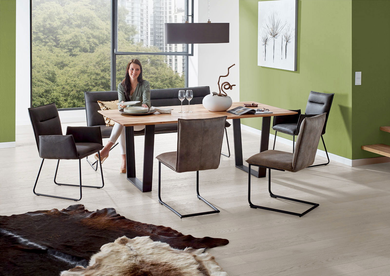 Valnatura Bank SCALA Moderner Essbereich mit Esstisch aus Holz und Metallgestell, umgeben von Freischwinger-Stühlen und einer Bank, in einem hellen Raum mit großen Fenstern und grüner Wand. Perspektive: seitlich.