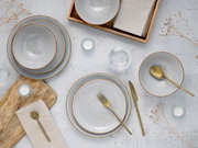 Minimalistisches Tischgedeck mit gesprenkeltem weißem Keramikgeschirr mit braunem Rand, goldfarbenem Besteck, Leinenservietten und Holztablett auf heller Steinoptik, Draufsicht (von oben)