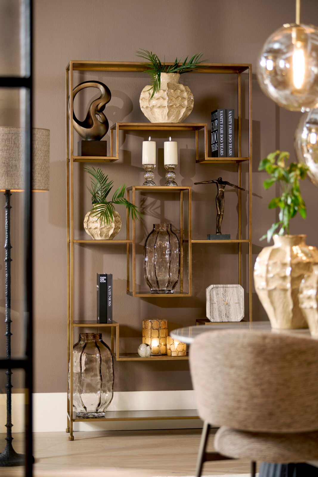 Vooraanzicht van een modern goudkleurig metalen wandrek met geometrische vakken, stijlvol gedecoreerd met vazen, kaarsen, boeken, planten en sculpturen in een warm interieur.