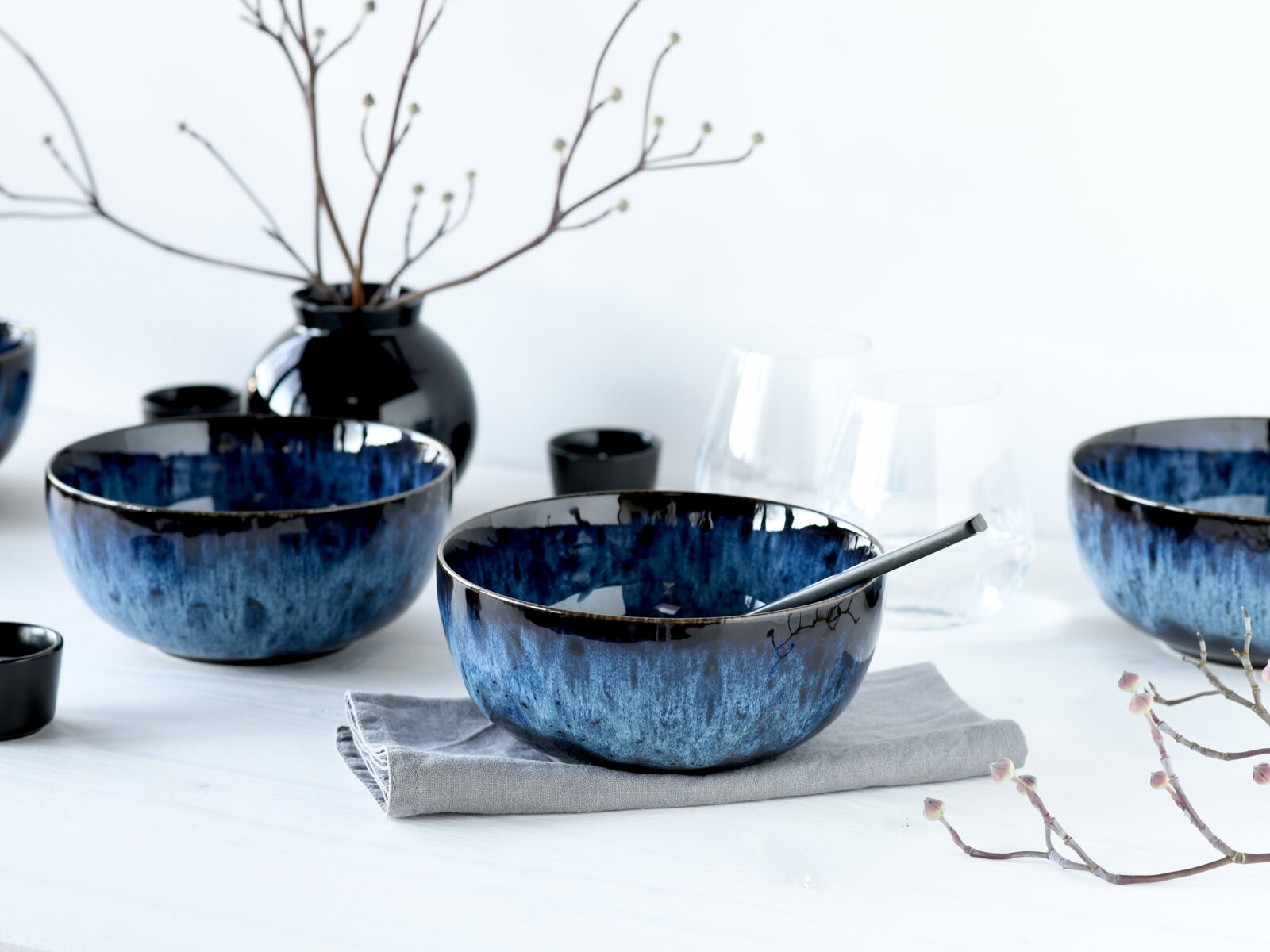 Blau glasierte Keramikschüssel mit dunklem Rand und Löffel auf grauem Leinentuch, umgeben von weiteren Schalen, Gläsern und schwarzer Vase auf hellem Tisch, aufgenommen in leichter Seitenansicht auf Tischhöhe.