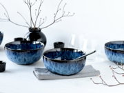 Blau glasierte Keramikschüssel mit dunklem Rand und Löffel auf grauem Leinentuch, umgeben von weiteren Schalen, Gläsern und schwarzer Vase auf hellem Tisch, aufgenommen in leichter Seitenansicht auf Tischhöhe.