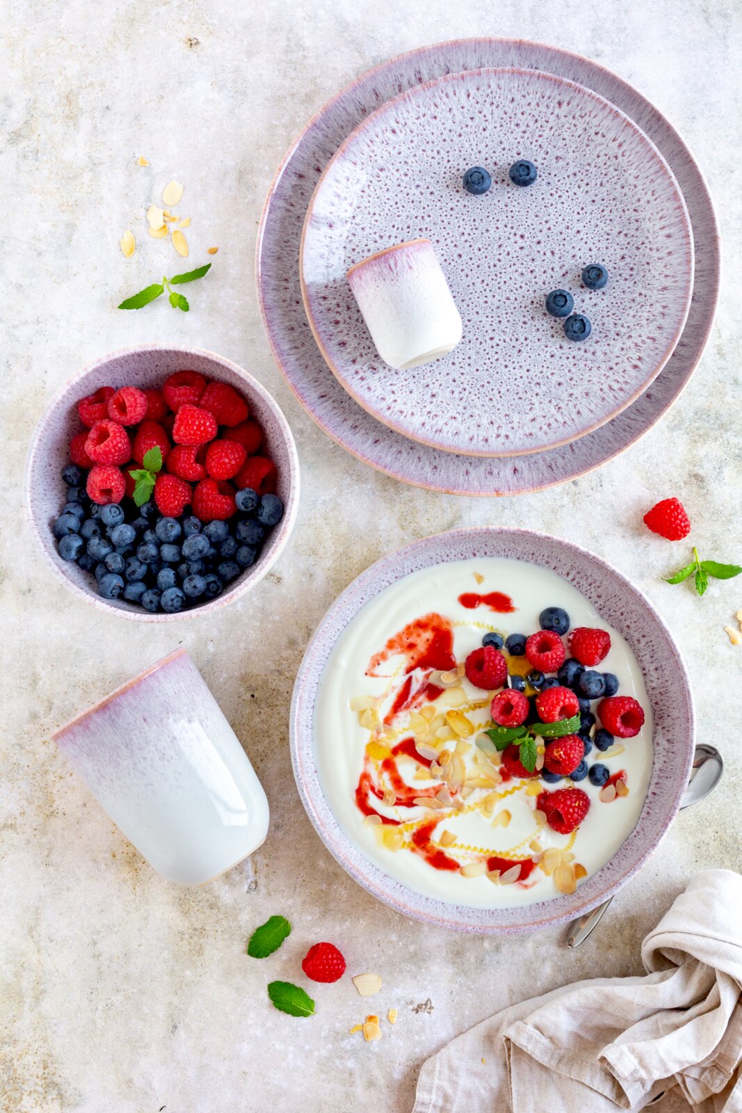 Keramik-Geschirrset in Rosa-Weiß mit Sprenkelglasur: großer Teller, Dessertteller, Schale und Becher, arrangiert mit Joghurt, Himbeeren, Blaubeeren, Mandeln und Minze; Draufsicht von oben.