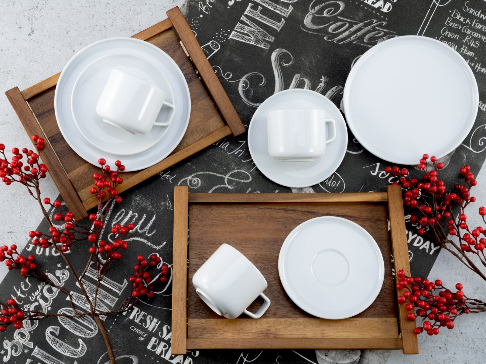 Draufsicht (Flatlay) auf ein weißes Porzellan-Geschirrset mit Tassen, Untertassen und Tellern auf Holztabletts, dekoriert mit roten Beerenzweigen auf einem schwarzen Coffee-Schrift-Hintergrund.