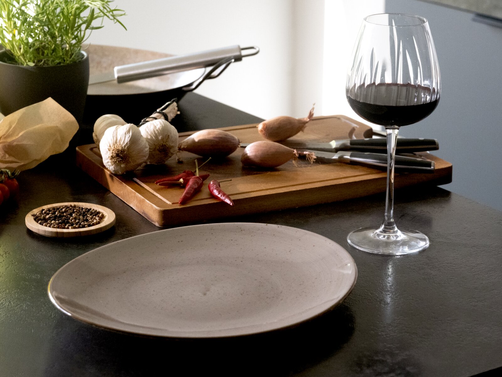 Rotweinglas mit Rotwein auf dunkler Küchenarbeitsplatte neben Holzschneidebrett mit Knoblauch, Schalotten und roten Chilis; Perspektive: leicht seitliche Ansicht auf Tischhöhe