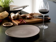 Rotweinglas mit Rotwein auf dunkler Küchenarbeitsplatte neben Holzschneidebrett mit Knoblauch, Schalotten und roten Chilis; Perspektive: leicht seitliche Ansicht auf Tischhöhe