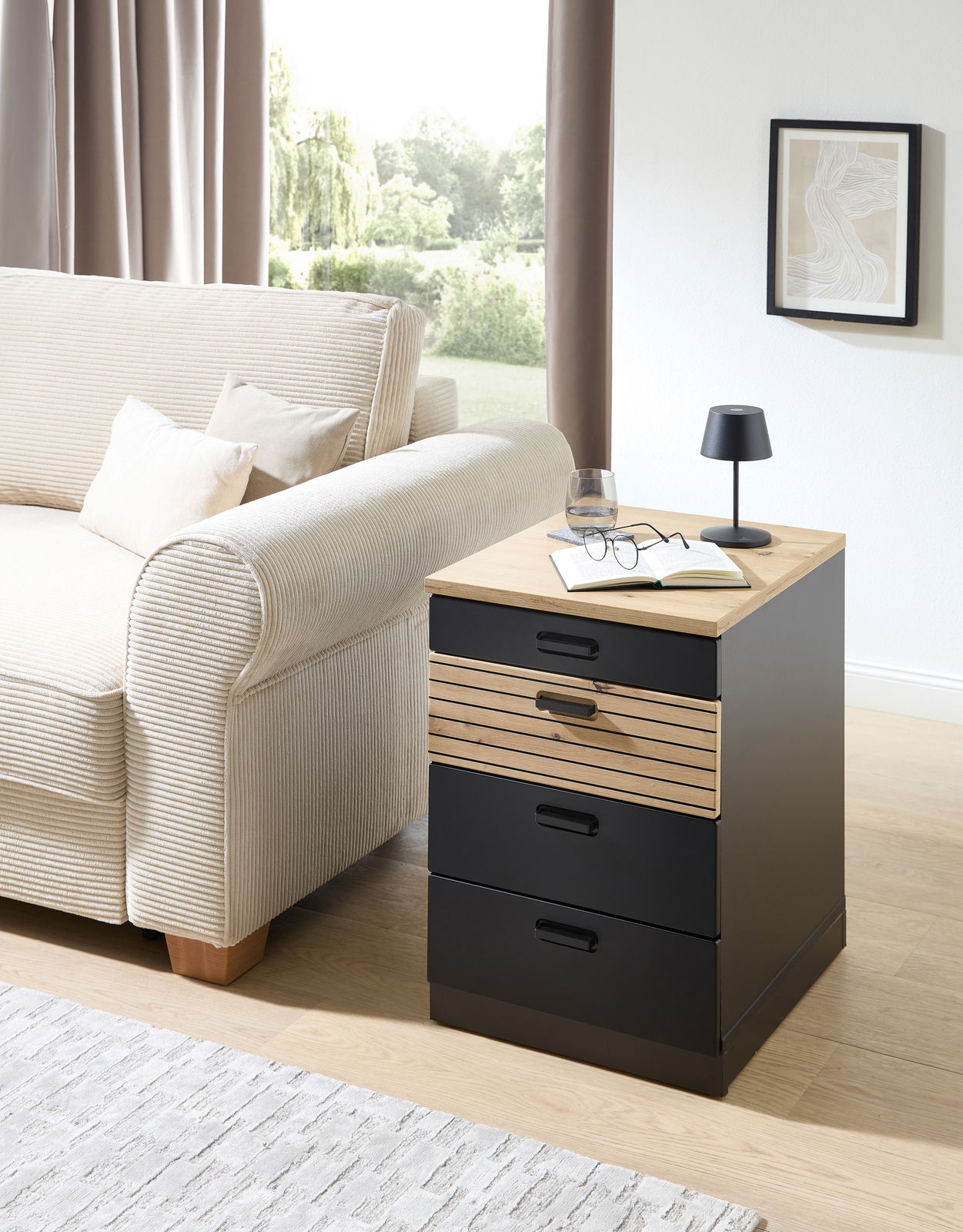 Rollcontainer mit drei Schubladen, aus schwarzem Material mit einer Holzoberfläche, neben einem Sofa in einem Wohnzimmer platziert. Perspektive von vorne rechts.