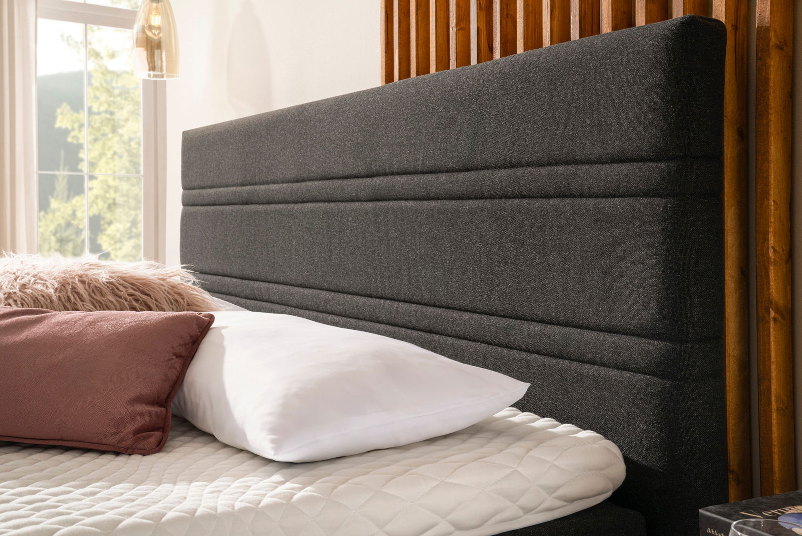 Nahaufnahme eines Boxspringbettes mit grauem Kopfteil, weißen und rosa Kissen, seitliche Perspektive.