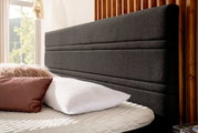 Nahaufnahme eines Boxspringbettes mit grauem Kopfteil, weißen und rosa Kissen, seitliche Perspektive.
