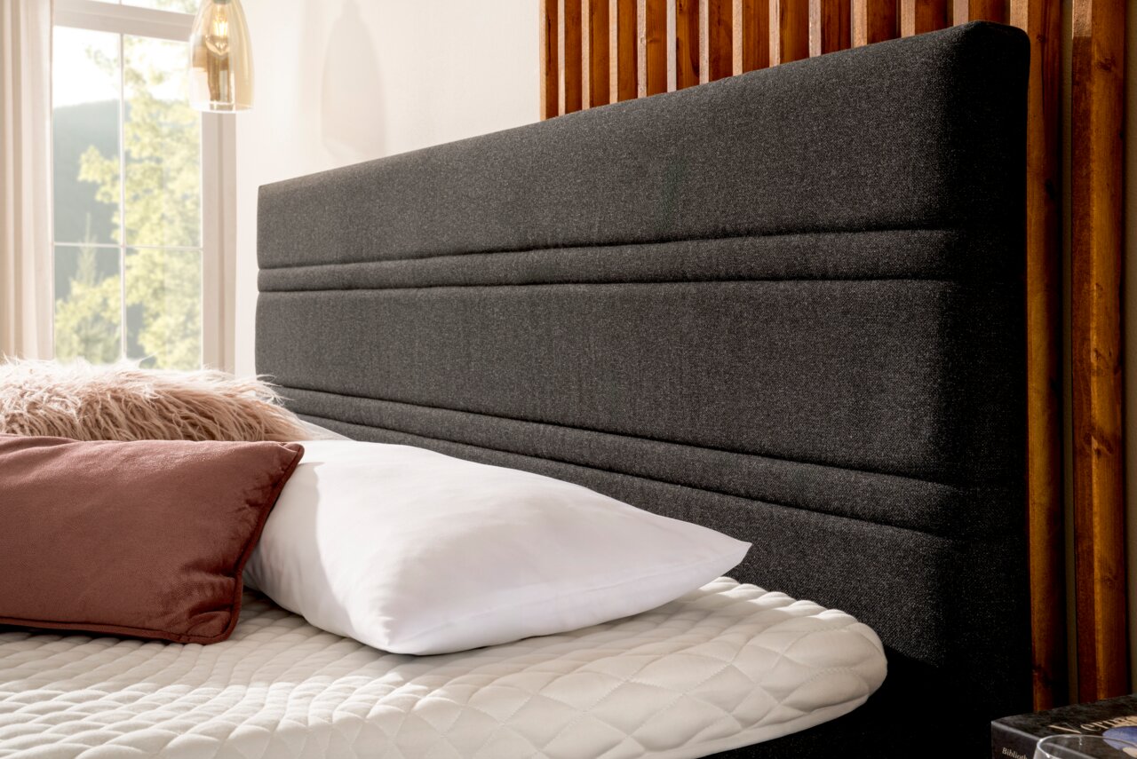 Nahaufnahme eines Boxspringbettes mit grauem Kopfteil, weißen und rosa Kissen, seitliche Perspektive.
