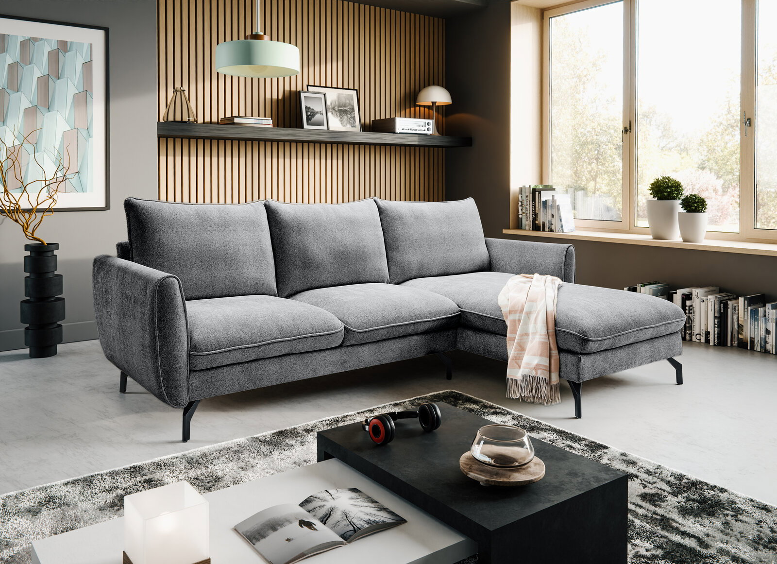 Modernes graues Ecksofa mit rechter Ottomane und schwarzen Metallfüßen in einem hellen Wohnzimmer, fotografiert in frontaler leicht schräger Perspektive.