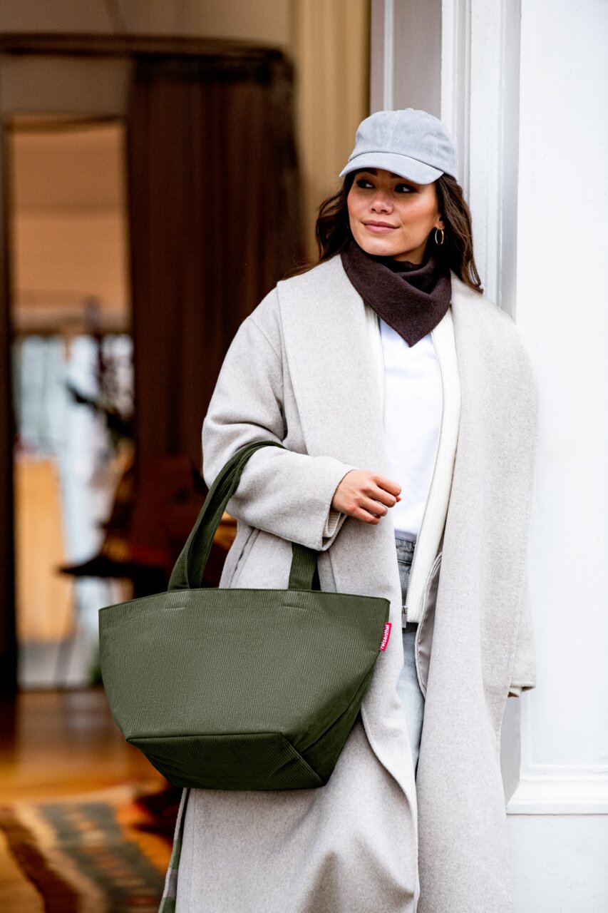 Olivgrüne Shopper-Tasche mit breiten Tragehenkeln, getragen am Unterarm einer Frau im hellen Mantel; Perspektive: frontale Halbtotalen-Ansicht leicht von links.