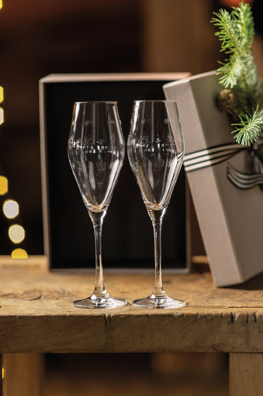 Zwei elegante Sektkelche auf einem Holztisch, im Hintergrund eine Geschenkbox und festliche Dekoration, seitliche Perspektive.