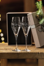 Zwei elegante Sektkelche auf einem Holztisch, im Hintergrund eine Geschenkbox und festliche Dekoration, seitliche Perspektive.